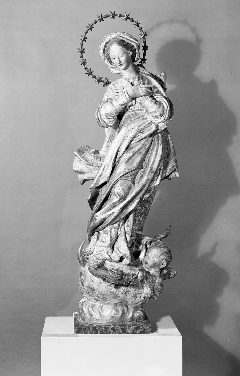 Wood statue of a standing Virgin Mary with a circle of stars around her head, arms crossed over her chest, robes draping off to her right, supported by a baby angel in a star-covered swaddle at the base.