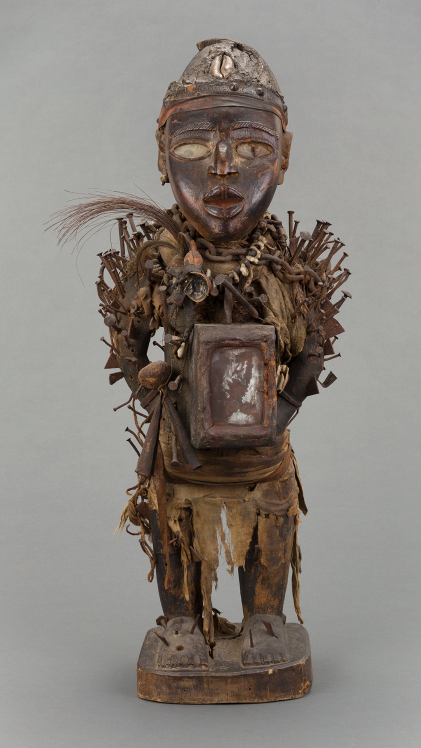 A wooden carving of a male figure. The figure has a distinctive face, with oval-shaped eyes and mouth, and wears a cap on his head. His body is covered in various shells, bones, feathers, fabric, and metal nails, and he holds a mirror-sealed resin packet over his stomach.