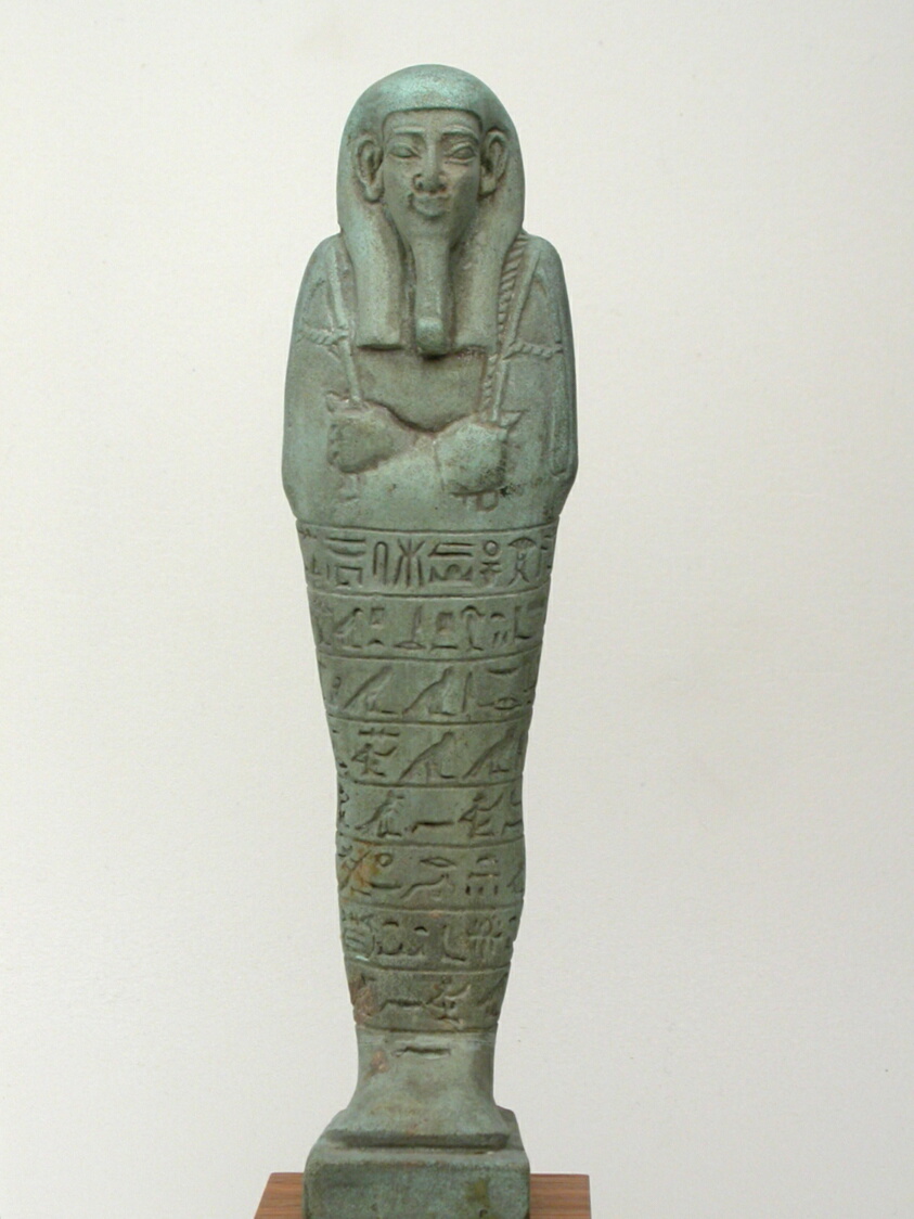 Green-gray sculpture of a mummy with hieroglyphs etched from below the crossed arms to just above the feet.