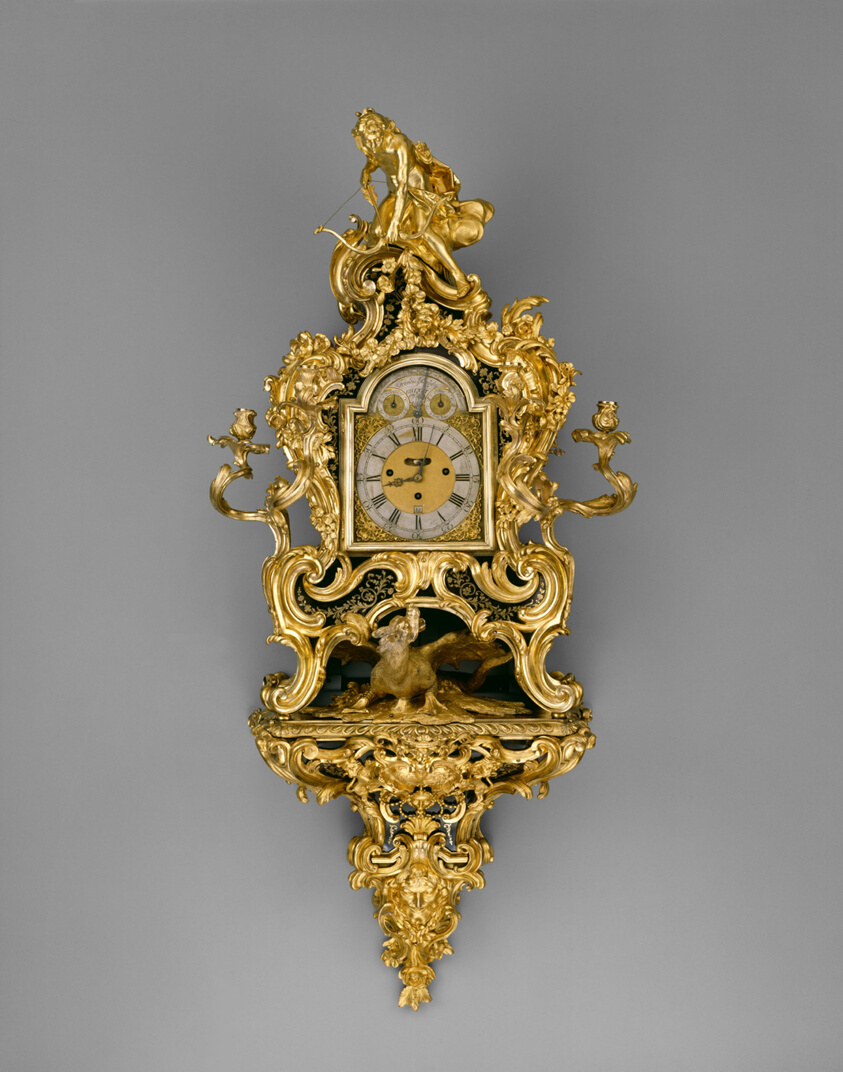 Golden-colored, ornate wall clock adorned with golden flourishes, a bent figure with an arrow at top.