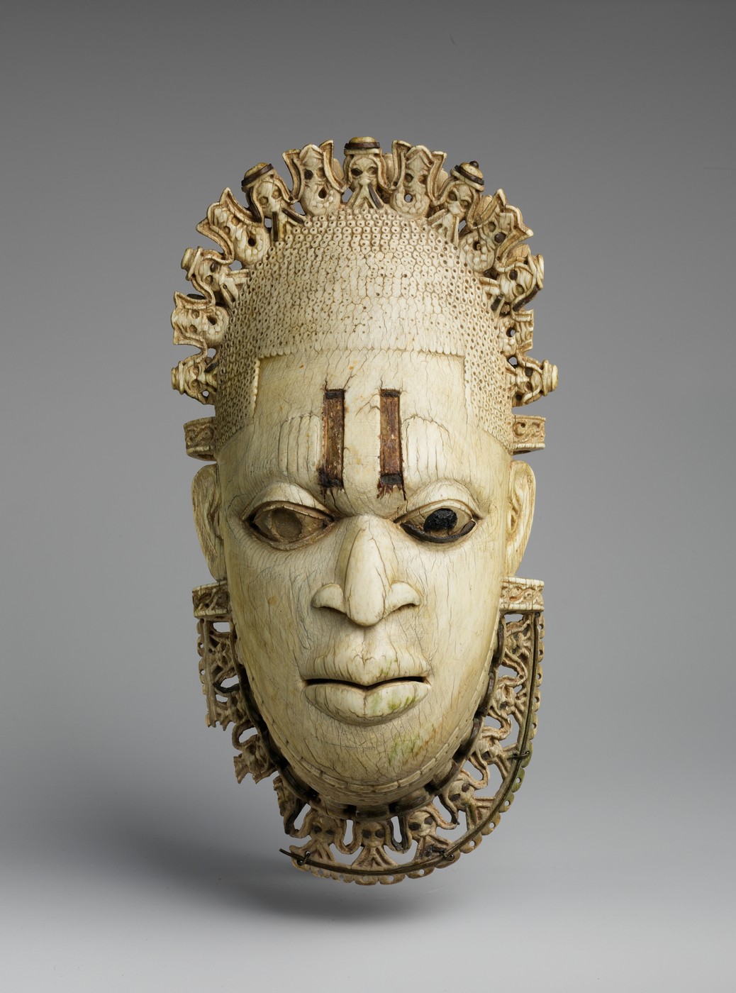 An ivory pendant mask with a finely carved face and an elaborate border; the Met example, not the British Museum FESTAC source.